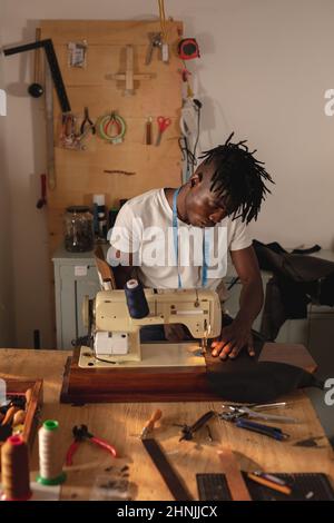 afroamerikanischer junger Handwerker, der während der Arbeit in der Werkstatt Leder auf Nähmaschine steppte Stockfoto