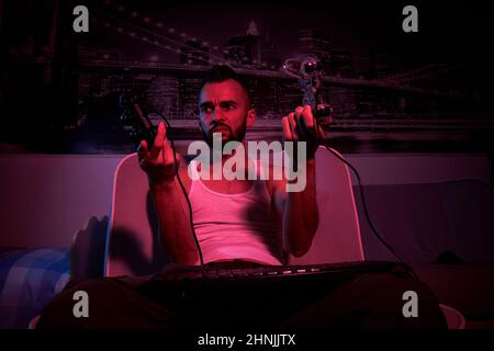 Ein 30-jähriger Mann mit einem Joystick in den Händen spielt Computerspiele, vergleicht die Joysticks, zeigt sie. Blick von der TV-Seite. Stockfoto