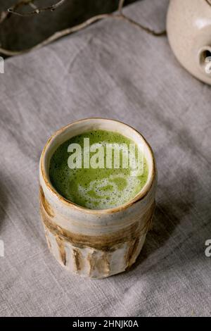 Nahaufnahme der traditionellen japanischen heißen grünen schaumigen Tee Matcha Latte in handgefertigter Keramik-Tasse, die auf einer grauen Leinentischdecke steht. Gesundes Getränk Stockfoto