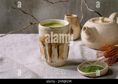 Traditioneller japanischer heißer grüner Milchtee-Matcha-Latte aus handgefertigter Keramik-Tasse, gepuderter Matcha, Teekannen und Bambusbesen auf grauem Leinentisch Stockfoto