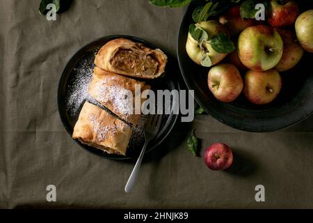 Hausgemachter, in Scheiben geschnittener traditioneller Apfelstrudelkuchen in schwarzer Keramikplatte, serviert mit reifen frischen Äpfeln, Ästen, Gabel und Puderzucker auf dunklem Leinentisch Stockfoto