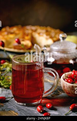 Hagebutten Beeren Kräutertee in Glas Becher stehen auf alten Holzbrett Tisch mit wilden Herbstbeeren und Apfelkuchen herum. Gesundes heißes Getränk. Stockfoto