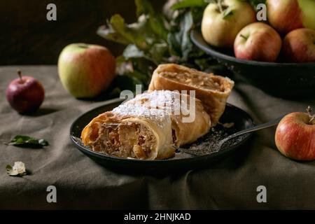 Hausgemachter klassischer Apfelstrudelkuchen in Scheiben in schwarzer Keramikplatte serviert mit reifen frischen Äpfeln, Ästen, Gabel und Puderzucker auf dunklem Leinentischlot Stockfoto