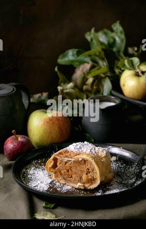 Scheibe hausgemachter traditioneller Apfelstrudelkuchen in schwarzer Keramikplatte, serviert mit reifen frischen Äpfeln, Zweigen, Gabel und Creme auf dunklem Leinentischtuch Stockfoto