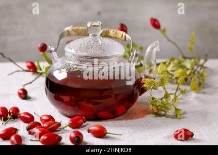 Hagebutten Beeren Kräutertee in Glas Teekane auf weißem Leinen Tischtuch mit wilden Herbstbeeren herum stehen. Gesundes heißes Getränk. Stockfoto