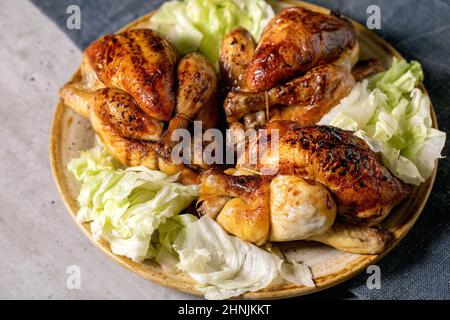 Drei ganze gebratene Mini-Hähnchen mit grünem Salat auf Keramikplatte auf blauer Leinentischdecke. Nahaufnahme Stockfoto