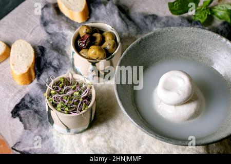 Traditioneller italienischer Burrata-Knüpfkäse in grauer Keramikschale auf dem Tisch. Brot, Oliven, grüner Salat. Zutaten für gesunde mediterrane din Stockfoto