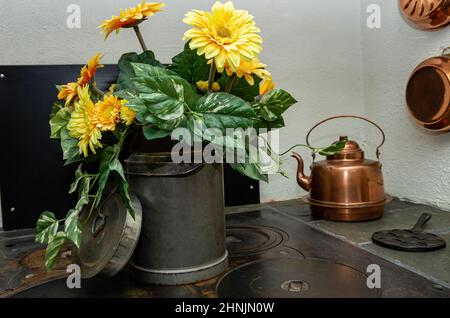 Ein alter Milchkännchen voller künstlicher Blumen wird zur Dekoration auf einen traditionellen Küchenherd gestellt. Stockfoto