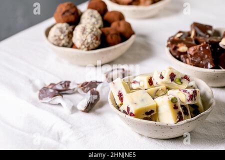 Auswahl an hausgemachten Schokoladen-Mandelnüssen aus Toffee, dunkler Schokoladentrüffel, weißer Schokoladen-Schokoriegel in Schüsseln auf weißem Tischtuch. Handgemachtes Essen Stockfoto