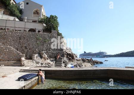 Das junge Paar genießt den Sommernachmittag am Strand von St. Jacob an der Adriaküste, während sich das Kreuzschiff Norwegian Jade dem Ankerplatz in der Lapad-Bucht von Dubrovnik nähert. Stockfoto