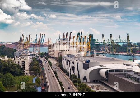Der riesige verkehrsreichste Logistikhafen in Singapur, viele Kräne zum Transport von Containern, riesige Frachtschiffe im Hintergrund, Einkaufszentren und Wolkenkratzer Stockfoto
