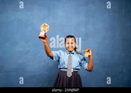 Genießen indische Schulmädchen Kind feiern, indem sie Trophäe und Medaille durch Blick auf Kamera - Konzept der Leistung, stolz, erfolgreich. Stockfoto