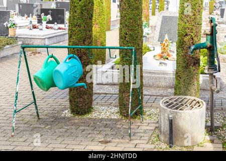 Gießkannen hängen neben einer alten Wasserpumpe auf dem städtischen Friedhof. Eine Handvoll Gartengeräte auf dem Friedhof, um sich um die Grünpflanzen zu kümmern Stockfoto