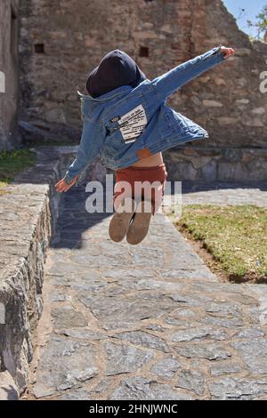 Kleiner Junge auf dem Rücken in einer Jacke mit Kapuze, die im Freien in der Luft springt. Kinderparkkonzept. Stockfoto