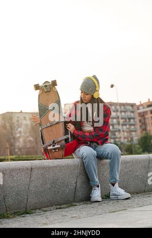 Junge Latina sitzt mit einem Rucksack und entfesselt das Skateboard. Venezolanische hispanische Frau, die Sport praktiziert. Stockfoto