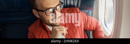 Ein hübscher kaukasischer Mann in legerer Kleidung und eine Brille, die nachdenklich aussieht, sitzt auf dem Flugzeug in der Nähe des Fensters Stockfoto