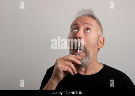 Mann mittleren Alters mit grauem Haar und Bart, der das Coronavirus (covid-19)-Antigen-Schnelltest-Kit (atk) zu Hause mit einem Nasenabstrich aus Baumwolle verwendet Stockfoto