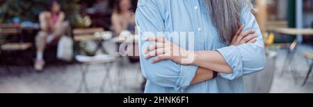 Positive haarige asiatische Dame mit gekreuzten Armen gegen die Terrasse des Cafés im Freien Stockfoto