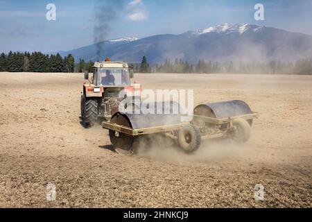 Tractor Pulling heavy metal Walze auf trockenen feld, Staub hinter und schwarzer Rauch oben, Berge im Rücken. Frühling Boden Vorbereitung. Stockfoto