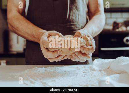 Die Hände des Küchenchefs sprühen Mehl über den Teig. Teig kneten. Der Chefkoch in der Schürze des Küchenchefs spritzt Mehl über den Teig Stockfoto