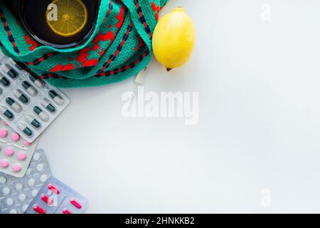 Eine Tasse Tee mit Zitrone steht auf einem gestrickten grünen und roten Schal, daneben befindet sich ein Thermometer, eine Zitrone und Pillen an der Seite. Stockfoto