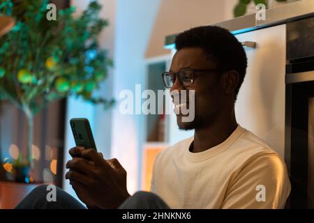 Junger glücklicher schwarzer Kerl mit dem Smartphone, während er sich entspannt und Freizeit im gemütlichen Zuhause verbringt Stockfoto