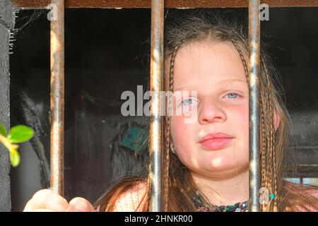 Foto eines kleinen Mädchens, das Bars hält. Trauriges kleines Mädchen hinter Eisenstangen. Kleines Mädchen hinter den Bars. Gewaltkonzept Stockfoto