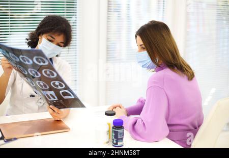 Arzt und Patient in schützenden Gesichtsmasken besprechen das Röntgenergebnis während des Termins. Afroamerikanische Ärztin mit CT-Lungenscreening Explainin Stockfoto