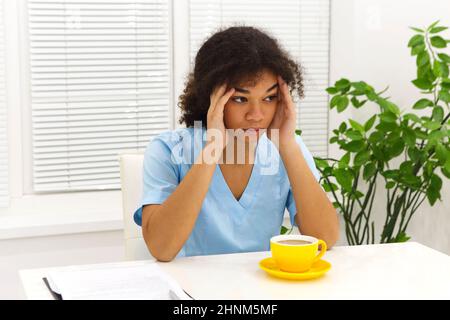 Müde depressive afroamerikanische Ärztin oder Krankenschwester in Uniform leiden nach hartem Arbeitstag oder schlaflosen Nachtschicht in der Klinik, sitt Stockfoto