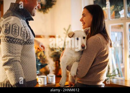 Junge glückliche Paar Mann und Frau mit niedlichen weißen Hund und umarmt, während in der Küche zu Weihnachten dekoriert stehen, schöne Familie mit Welpen enjoyi Stockfoto
