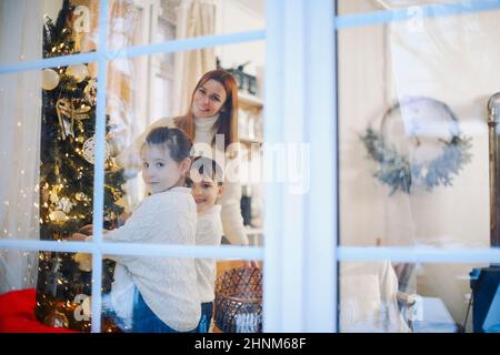 Süße kleine Kinder helfen Eltern, Weihnachtsbaum zu Hause schmücken, glückliche Eltern und kleine Kinder, die sich auf Weihnachten und Neujahr vorbereiten, schmücken hous Stockfoto