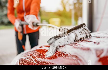 Junge Frau, die ihr Auto in einer Autowäsche mit Selbstbedienung putzt. Nahaufnahme Detail auf weißem Seifenschaum und Bürste über Dach bewegen. Stockfoto