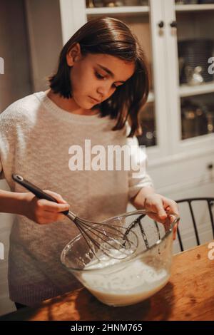 Fröhliches Teenager-Mädchen, das in der Küche kocht, lächelndes kleines Mädchen, das hinter dem Tisch steht und Eier und Milch in einer Schüssel hastet, während es Teig für hausgemachtes cak zubereitet Stockfoto