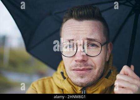 Porträt eines jungen Mannes mit Brille unter Regenschirm im Regen. Themen Wetter eine Meteorologie. Stockfoto