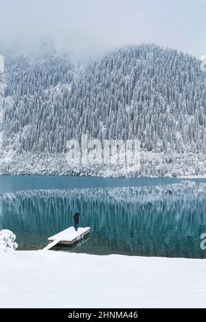 Im Winter steht ein Mann auf dem Bootssteg eines Bergsees Stockfoto