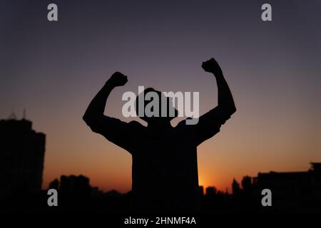 Silhouette männlich tanzen und glücklich fühlen im Freien bei Sonnenuntergang mit städtischem Hintergrund. Mann pumpt seine Fäuste in die Luft, Sieg Geschäftsmann Konzept, tanzen, die Hände heben und winken. Hochwertige Bilder Stockfoto