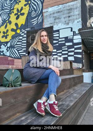 Im Herbst sitzt eine junge Frau auf einer Holztreppe in der Nähe eines Gebäudes in einem Künstlerviertel Stockfoto