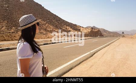 Ein weißes Mädchen in einem Hut steht in der Nähe einer breiten, menschenleeren Asphaltstraße. Rückportrait einer Frau, die auf einer leeren Straße mit Bergen und heißer Sonne in die Ferne blickt. Nahe Osten. Reisekonzept Stockfoto