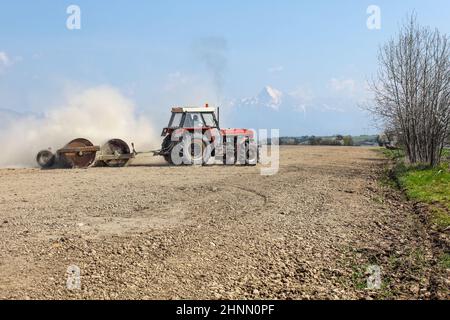 Liptovsky Hradok, Slowakei - 22nd. April 2018: Traktor zieht Schwermetallrolle, bereitet trockenes Feld im Frühjahr vor, Staubwolke dahinter, mit Berg Krivan, slowakisches Symbol, im Hintergrund. Stockfoto