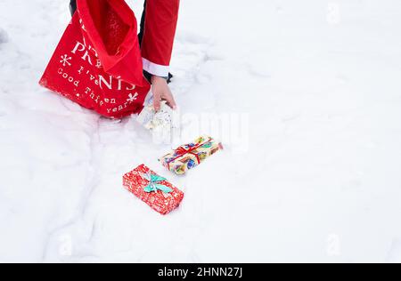 Ein Mann, der als Weihnachtsmann gekleidet ist, sammelt in einem verschneiten Wald im Schnee Geschenke, bunte Schachteln mit Geschenken. Stockfoto