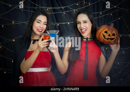 Zwei Frauen in rotem Kleid und schwarzem Mantel halten ein Cocktailglas und Kürbis in ein Gesicht geschnitzt genießen halloween-Party mit blinkenden Lichtern geschmückt. Stockfoto