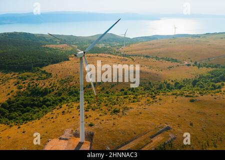 Windmühlen erzeugen grüne Energie an einer elektrischen Station auf einem mit Büschen bedeckten Hügel gegen Wälder in der Nähe des Adriatischen Meeres bei sonnigem Blick von der Drohne aus Stockfoto