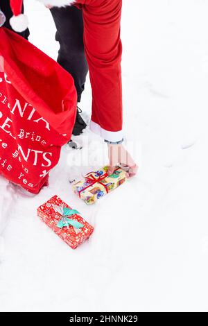 Ein Mann, der als Weihnachtsmann gekleidet ist, sammelt in einem verschneiten Wald im Schnee Geschenke, bunte Schachteln mit Geschenken. Stockfoto