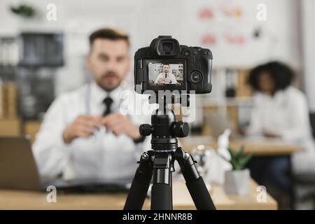 Kompetenter medizinischer Mitarbeiter in Uniform, der Live-Streaming macht, während er am Schrank sitzt. Fokus auf dem Bildschirm der Digitalkamera. Sendung zum Thema Medizin. Stockfoto