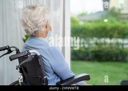 Eine ältere Frau, die auf einem Rollstuhl sitzt und aus dem Fenster schaut, um jemanden zu warten. Traurig, melmütig und deprimiert. Stockfoto