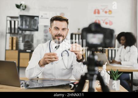 Kaukasischer Arzt sitzt am Schreibtisch mit elektronischem Thermometer in den Händen und zeichnet Videos auf professioneller Kamera auf. Telemedizin, Menschen- und Technologiekonzept. Stockfoto