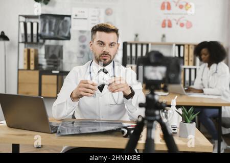 Kaukasischer Arzt sitzt am Schreibtisch mit elektronischem Thermometer in den Händen und zeichnet Videos auf professioneller Kamera auf. Telemedizin, Menschen- und Technologiekonzept. Stockfoto