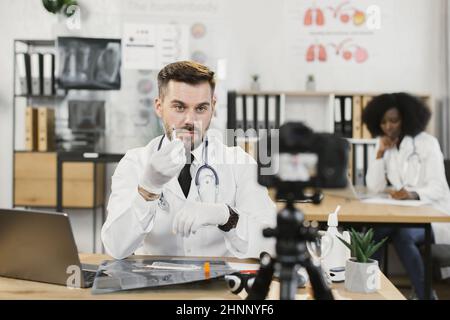 Qualifizierter männlicher Arzt in Laborkittel und Handschuhen mit steriler Spritze und Gespräch über Injektion während der Aufnahme Video-Blog. Kaukasischer Mann mit modernem Laptop und Kamera. Stockfoto