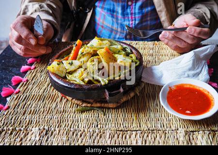 Tajine oder Tagine: Traditionelle Berberspeise, die in einer Steingutschüssel in einem typischen marokkanischen Straßenrestaurant serviert wird. Details aus der Nähe, man hält die Gabel und Messer im Hintergrund, während er gerade zu essen ist Stockfoto