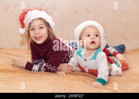 Porträt eines kleinen lächelnden Mädchens und Jungen in Pullovern und Weihnachtsmannmützen. Porträt auf beigefarbenem Hintergrund Stockfoto
